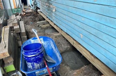 A blue bucket with a shovel on it next to a blue house undergoing foundation repair.