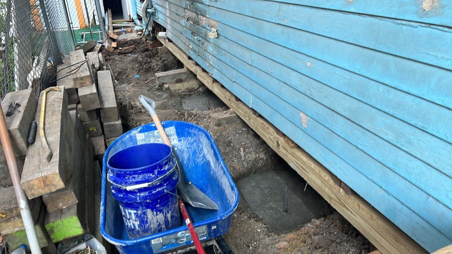 A blue bucket with a shovel on it next to a blue house undergoing foundation repair.