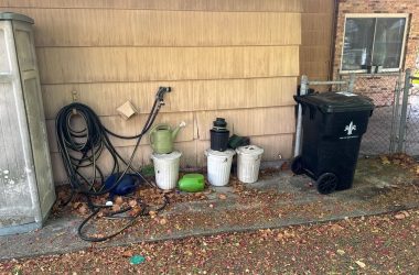 A black trash can sits outside of a house.