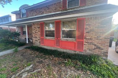 A brick house with red shutters and a car parked in front of it that requires foundation repair.
