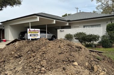A large pile of dirt is situated in the driveway of a residential home in the Southeast, with an "academy house leveling" sign posted, indicating foundation repair methods or leveling work in progress.