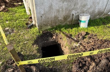 A caution tape surrounds an open excavation hole by the foundation of a building, essential for inspecting rotten pilings, with a bucket nearby.