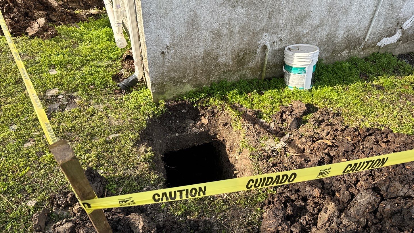 A caution tape surrounds an open excavation hole by the foundation of a building, essential for inspecting rotten pilings, with a bucket nearby.