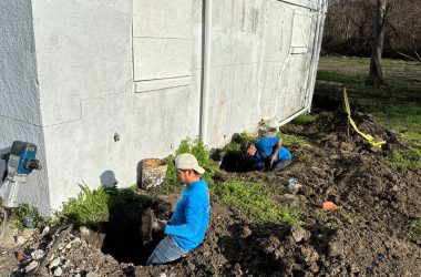 Two workers are digging a hole for drainage solutions near the exterior wall of a building on a sunny day.