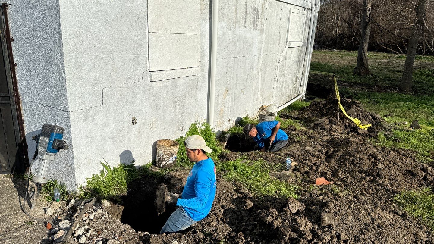Two workers are digging a hole for drainage solutions near the exterior wall of a building on a sunny day.