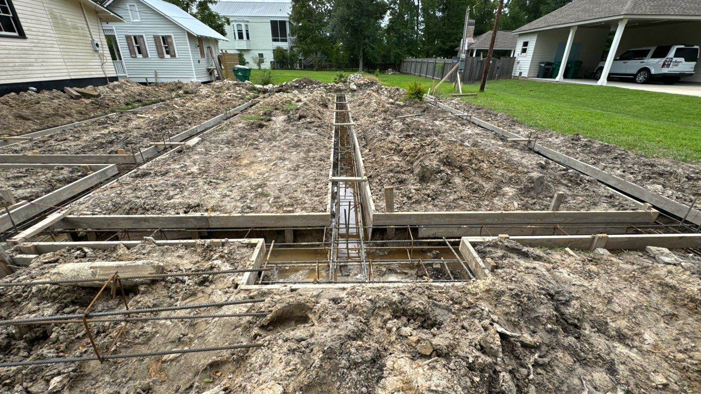 A construction site with foundation footings being prepared. Metal rebar and wooden forms are set in trenches, surrounded by dug-up soil. In the background are residential buildings and green grass, reminiscent of a bustling New Orleans neighborhood.