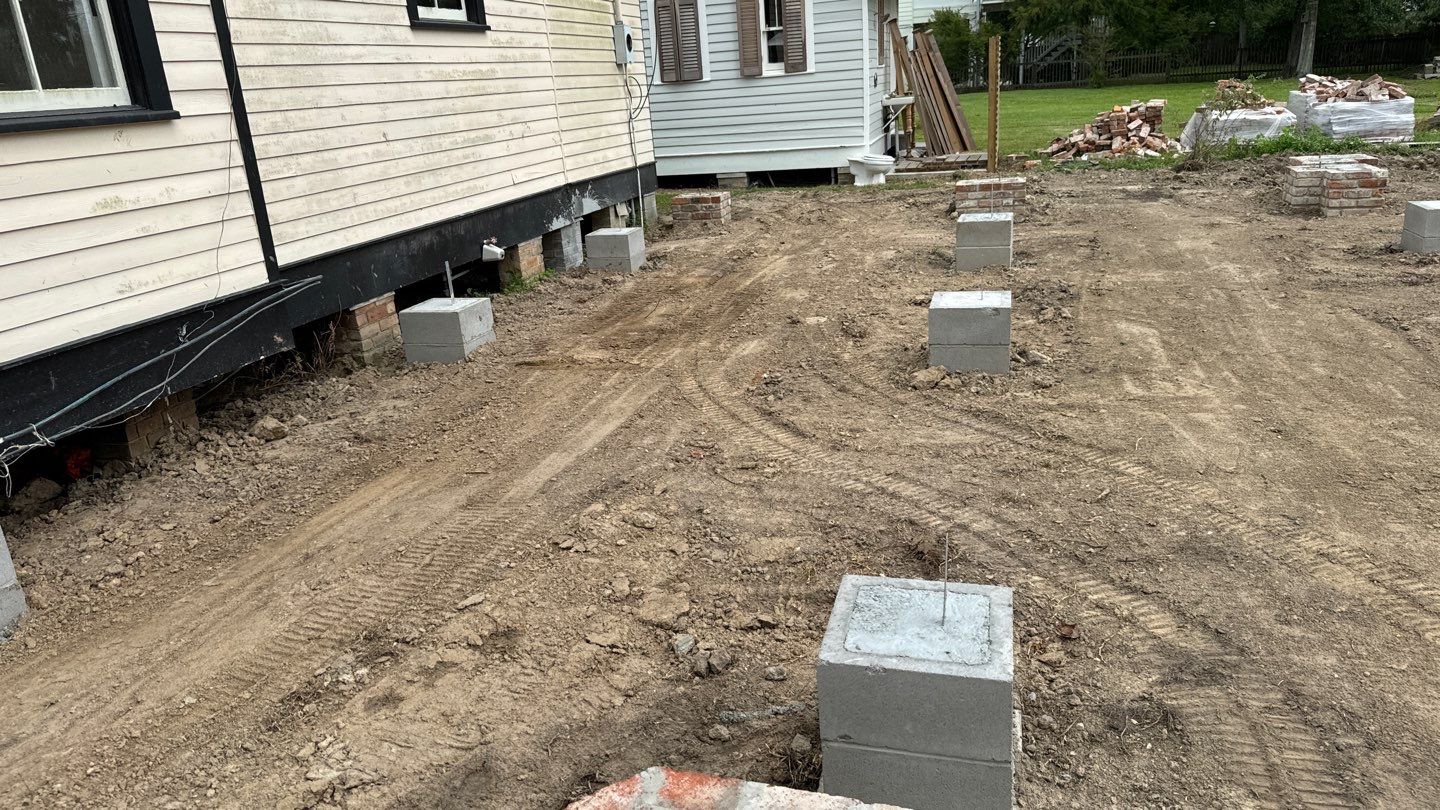 A house resting on concrete blocks with freshly cleared soil showing the layout and foundation footings. Brick piles and construction materials are visible in the background, ensuring regular inspections maintain progress and safety standards.