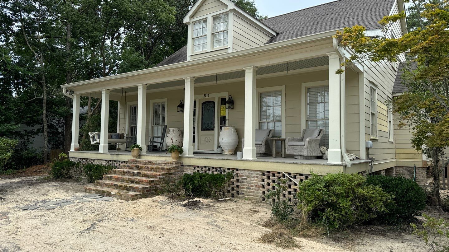 A beige house with a gabled roof and a front porch featuring white columns, wicker furniture, and potted plants. Located in Mandeville, the front yard is partially unpaved with sparse vegetation, reflecting a bit of that classic New Orleans charm.