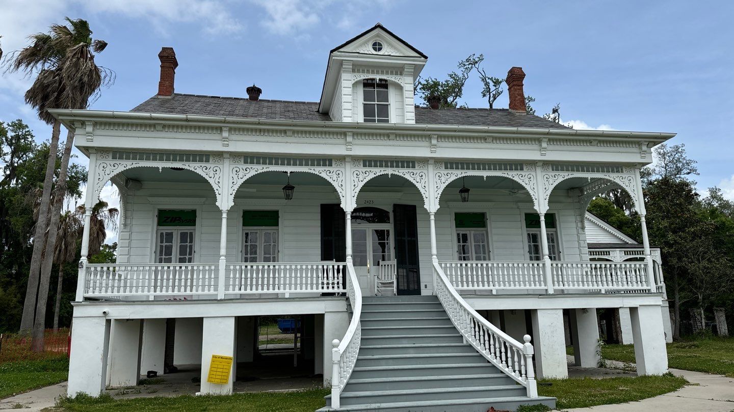 A two-story white house with intricate trim, elevated on stilts to avoid foundation issues. It features a large front porch and is nestled in Biloxi, MS, with a picturesque palm tree and other lush vegetation in the background.