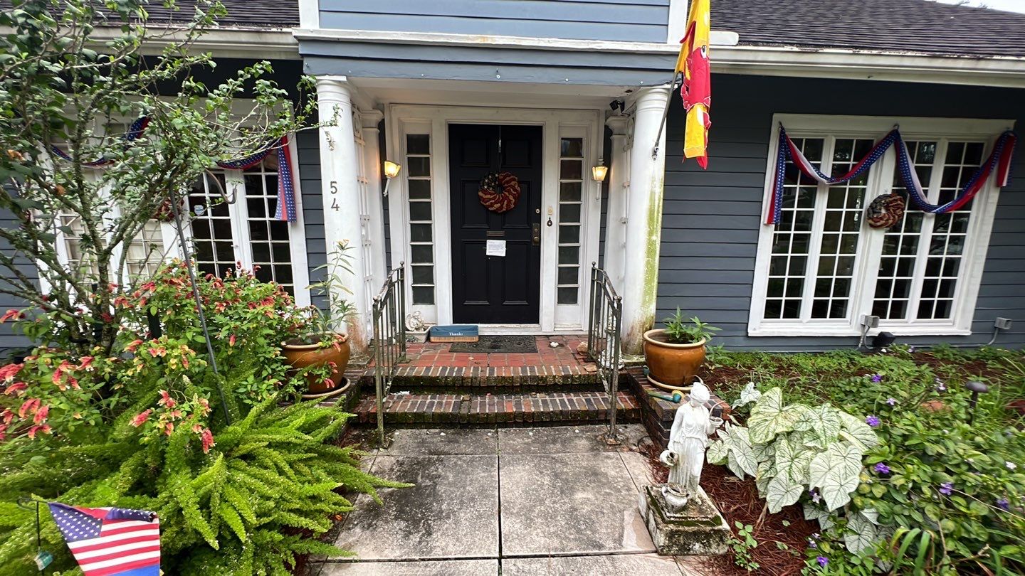 A blue house with a black door, flanked by two potted plants. The entrance features white columns and decorative greenery. An American flag and another flag hang near the door, making it an inviting sight for anyone home buying in New Orleans.