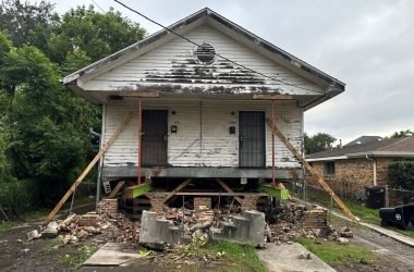 A small house in Jefferson Parish, elevated on wooden beams, shows wear and damage, with bricks scattered around its base and supports steadied on either side. House leveling might be needed to restore stability to this Metairie home.