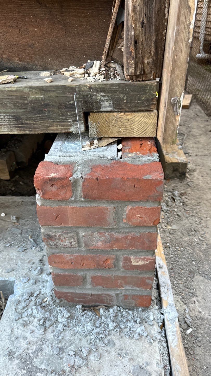 A small brick pillar supports a wooden beam, possibly part of a home elevation project in New Orleans. Wood and metal fragments are scattered on top of the beam, indicating active construction or renovation work.