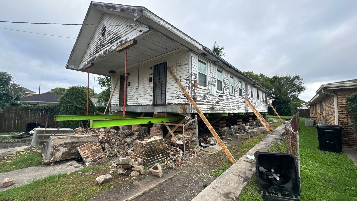 A weathered, single-story house in Metairie is elevated on temporary stilts with bricks and wooden supports beneath, as part of a house leveling project. The house shows signs of wear, with peeling white paint and a partially collapsed porch.