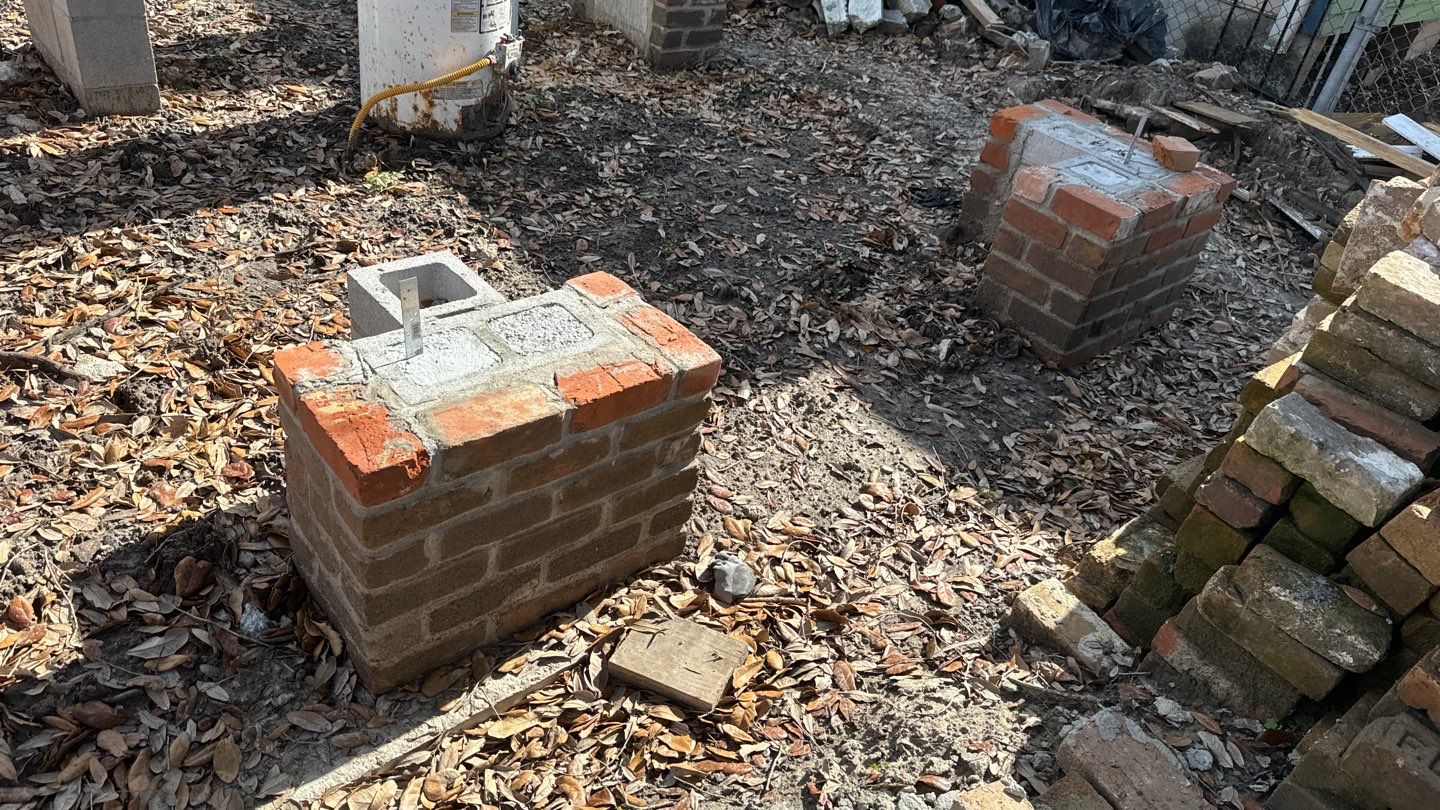 Two newly constructed brick pillars stand on a leaf-covered ground. A stack of bricks is nearby, along with various construction materials in the background—key elements for a home elevation project in New Orleans.