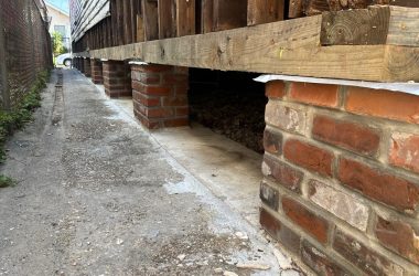 A building foundation supported by brick pillars, with a concrete pathway and metal fence on the left side, showcases the classic techniques of home elevation commonly seen in New Orleans.