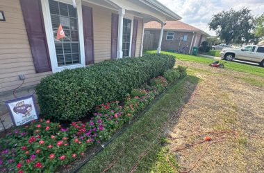 A well-maintained front yard showcasing trimmed bushes, blooming flowers, and a lawnmower nearby. The property in Harvey, Louisiana features a beige-house exterior with white-trimmed windows and dark shutters. Regular foundation inspections can help prevent common issues in properties like these.