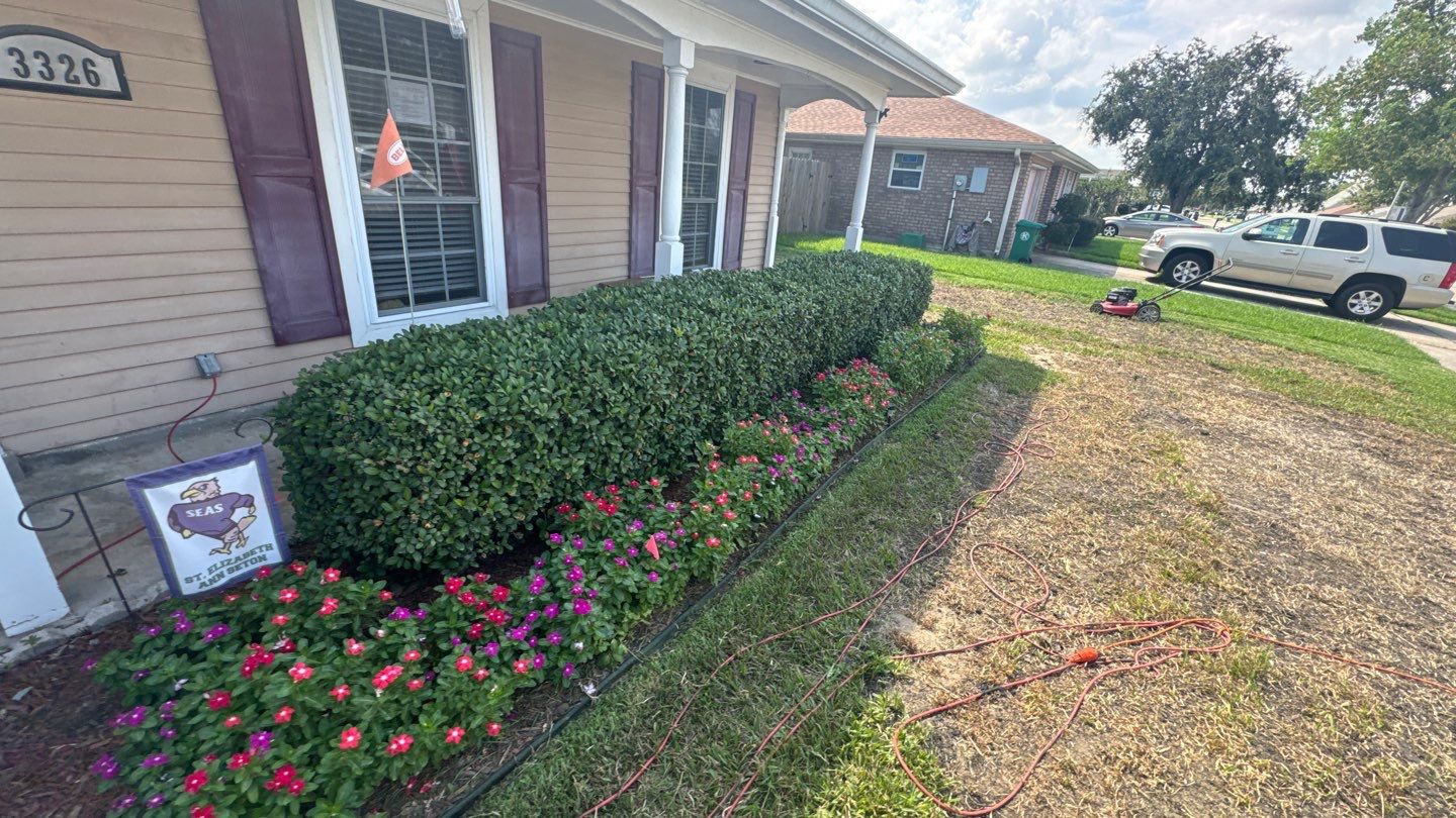 A well-maintained front yard showcasing trimmed bushes, blooming flowers, and a lawnmower nearby. The property in Harvey, Louisiana features a beige-house exterior with white-trimmed windows and dark shutters. Regular foundation inspections can help prevent common issues in properties like these.