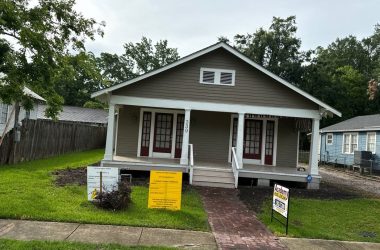 A single-story gray house with a front porch and two sets of French doors awaits its new residents. Located in charming New Orleans, the yard features a sidewalk leading to the porch and two signs near the walkway, including a "For Sale" sign. House leveling has been meticulously maintained.
