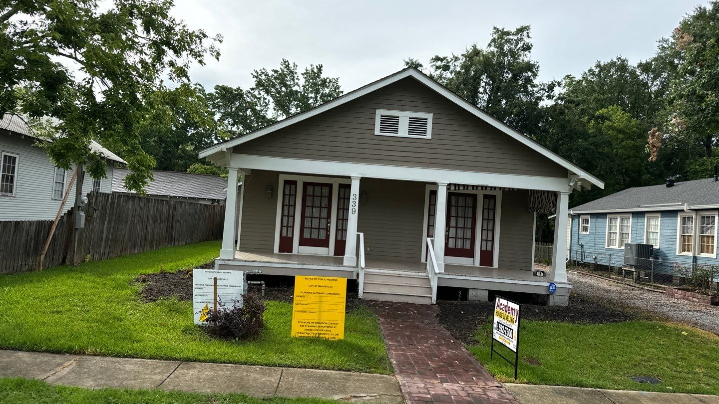 A single-story gray house with a front porch and two sets of French doors awaits its new residents. Located in charming New Orleans, the yard features a sidewalk leading to the porch and two signs near the walkway, including a "For Sale" sign. House leveling has been meticulously maintained.