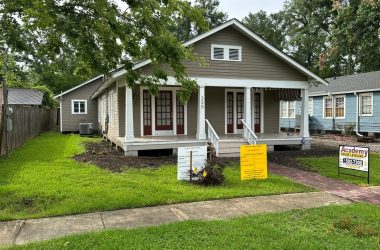 A one-story house with a front porch and steps, beige siding, and white accents. Multiple signs are in the front yard, including for sale and construction signs, indicating possible foundation repair. Neighboring houses are visible.