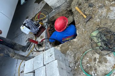 A construction worker wearing a red helmet works on the foundations inside a hole at a Kenner LA construction site, surrounded by tools, equipment, and bricks. Another worker stands nearby, ready to address any drainage issues that may arise.