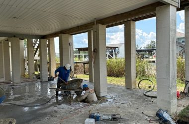 Three workers perform construction tasks in an open-sided building with columns in Bay St. Louis. One operates a wheelbarrow, another sits working on the ground, and a third person bends over near equipment, ensuring everything is perfect for upcoming yearly home inspections.