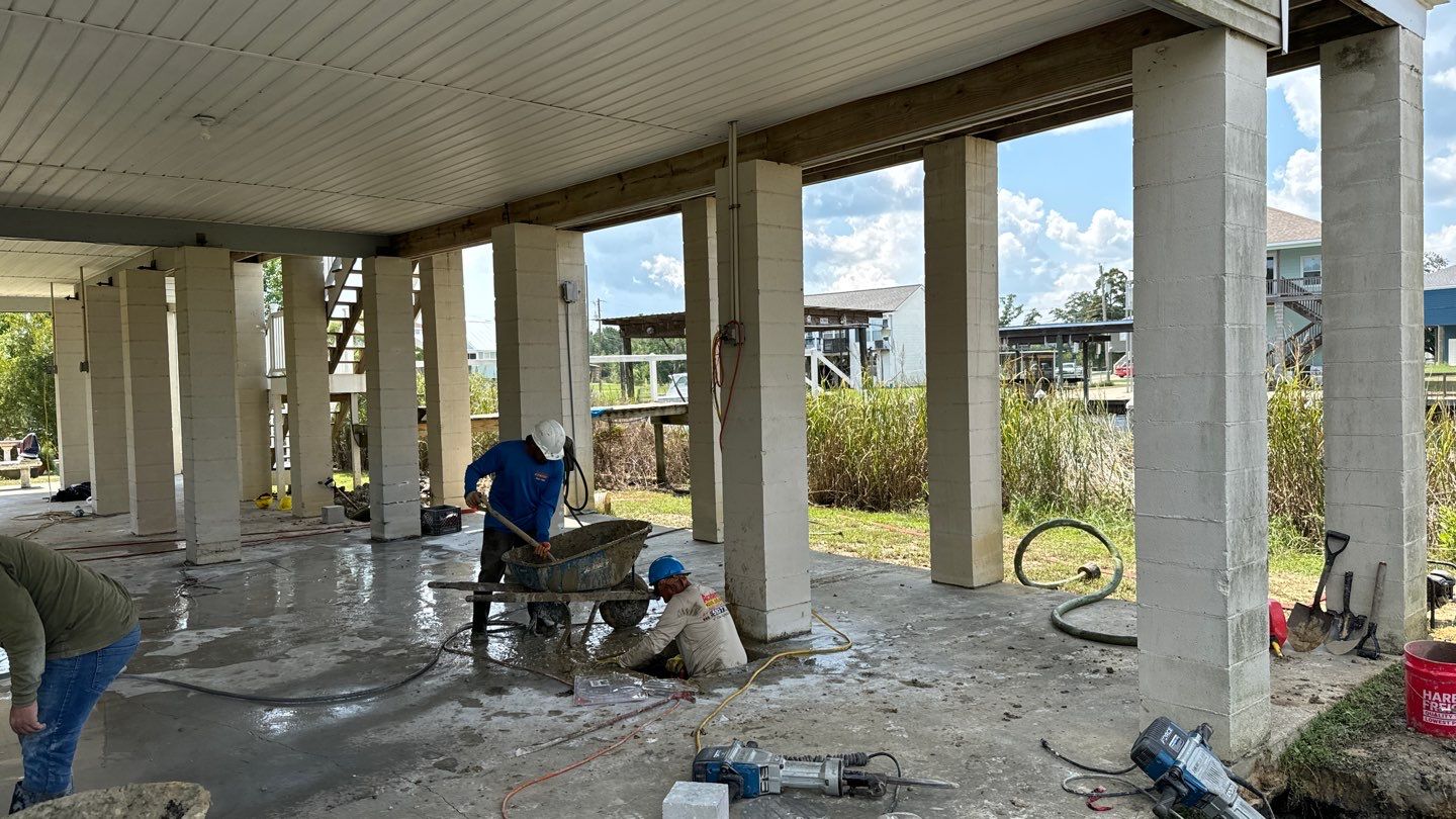 Three workers perform construction tasks in an open-sided building with columns in Bay St. Louis. One operates a wheelbarrow, another sits working on the ground, and a third person bends over near equipment, ensuring everything is perfect for upcoming yearly home inspections.