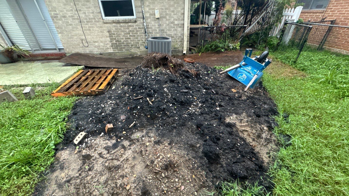 A pile of soil with scattered debris sits in a Louisiana backyard near a brick house, accompanied by gardening tools, a wooden pallet, and an overturned wheelbarrow—perfect for anyone ready to level a home or simply enjoy some leveling time.