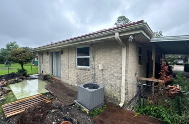 A small beige brick house in Louisiana, featuring a red roof and a metal awning, stands on a concrete patio surrounded by a wet yard. An air conditioning unit is nestled next to the home. The overcast sky adds to the charm of this level-a-home beauty.