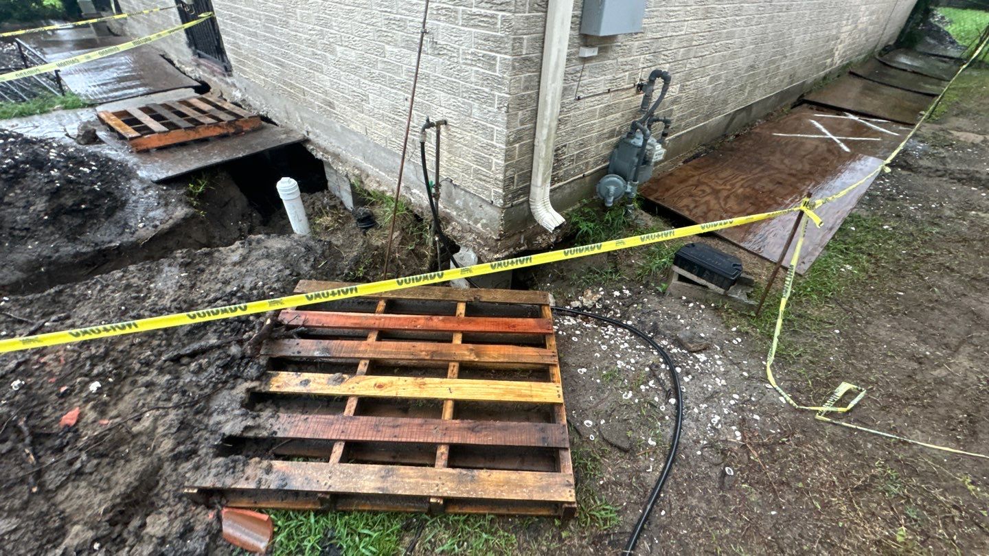 A section by a brick wall in Louisiana is cordoned off with yellow caution tape. Wooden pallets cover the muddy ground next to a partially visible house, hinting at ongoing efforts to level a home. Wet ground and scattered construction materials complete this temporary scene.