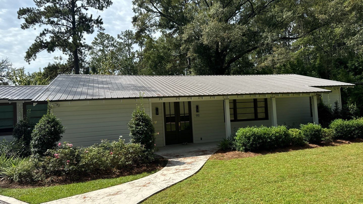 A single-story house with a metal roof, commonly found among homes in Louisiana, is surrounded by lush greenery and trees. A stone path leads to the front door, offering ample charm despite potential foundation issues.