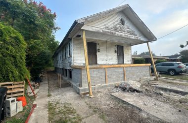 A single-story white house in New Orleans, set on a raised foundation, is undergoing home leveling and renovation. Piles of debris are scattered in front, with a couple of parked cars nearby.