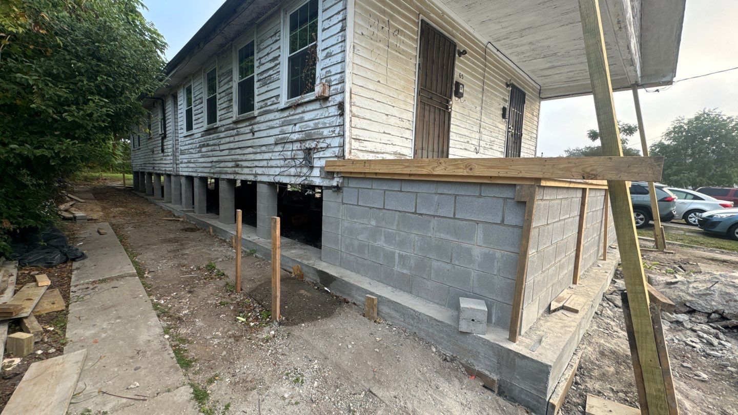 A house in New Orleans is undergoing renovation, featuring visible structural repairs and a new cinder block foundation. Home leveling work is underway, with scaffolding in place and construction materials scattered about.