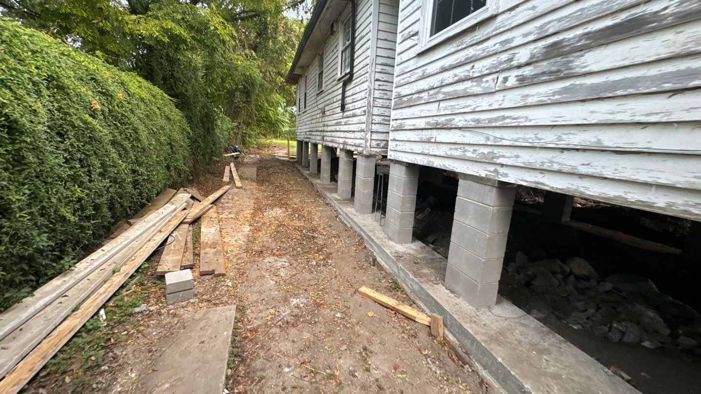 A house in New Orleans stands elevated on concrete blocks, its paint peeling. Wooden planks are stacked nearby, hinting at a project from the 2024 Home Leveling Guide. Tall trees line the property, offering shade and charm to this classic Southern abode.
