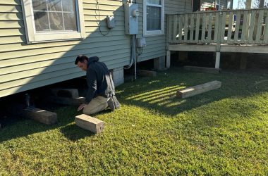A person kneels on the grass beside a house in Louisiana, carefully inspecting the pier and beam foundation.