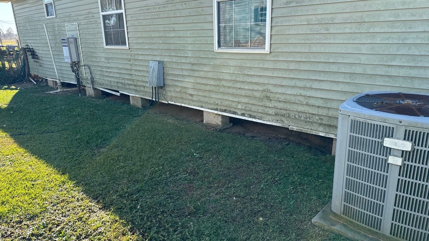 The side of the house features gray siding, windows, an air conditioning unit, and a grassy yard. In this Louisiana setting, the foundation shows signs of erosion beneath, suggesting a need for pier and beam foundation repair to maintain stability.
