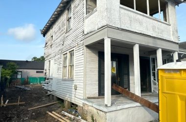 A two-story house in New Orleans is under renovation, showcasing peeling white paint and exposed wood. A ramp leads to the front entrance, hinting at updates to its pier and beam foundations. A yellow portable toilet sits beside the home, underscoring the importance of ongoing work.