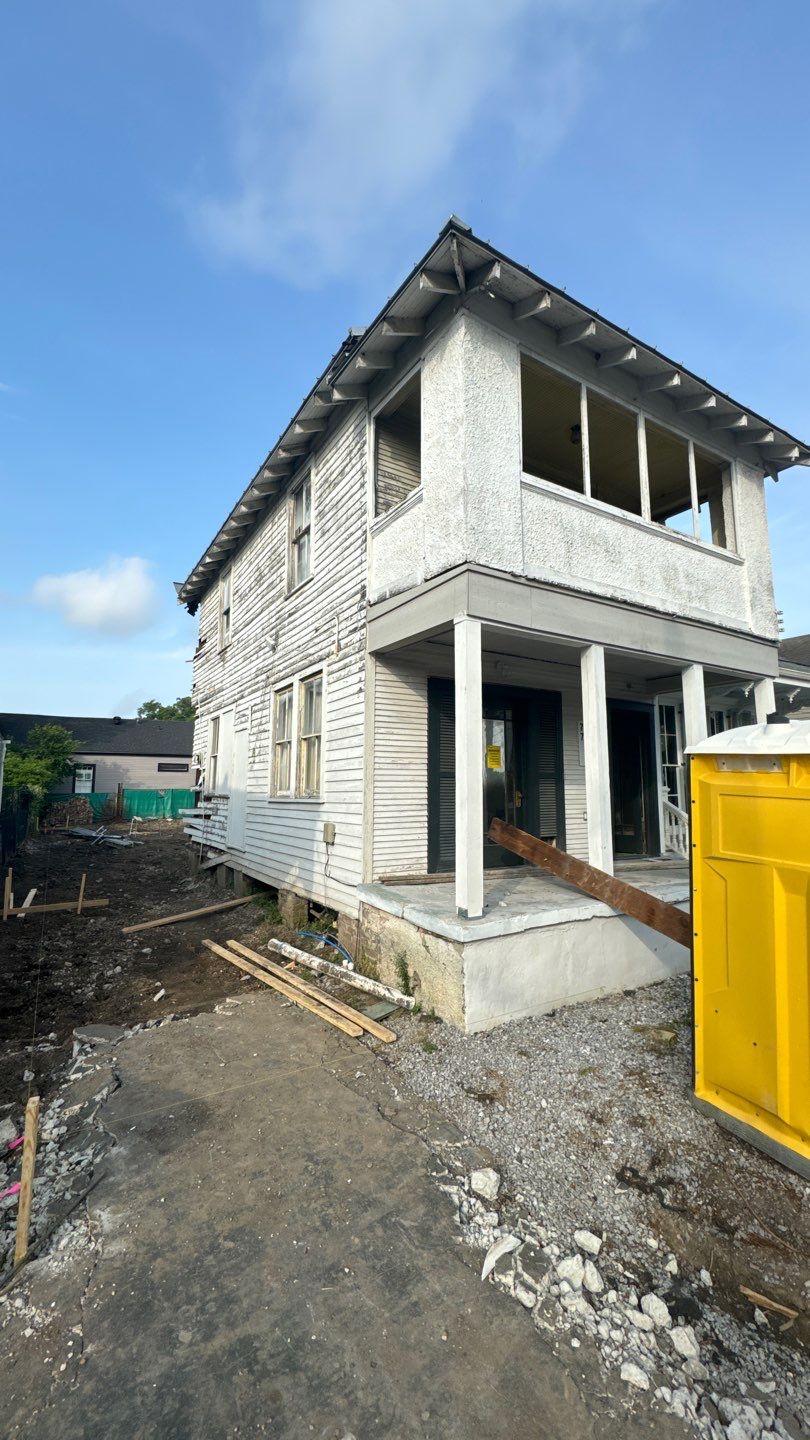 A two-story house in New Orleans is under renovation, showcasing peeling white paint and exposed wood. A ramp leads to the front entrance, hinting at updates to its pier and beam foundations. A yellow portable toilet sits beside the home, underscoring the importance of ongoing work.
