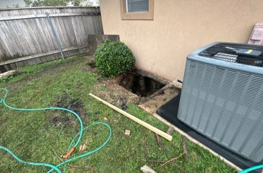 A small yard in Baton Rouge features a hole next to a house, surrounded by a bush, garden hose, bricks, and an air conditioning unit—an essential site for home leveling to ensure the foundation stays secure.