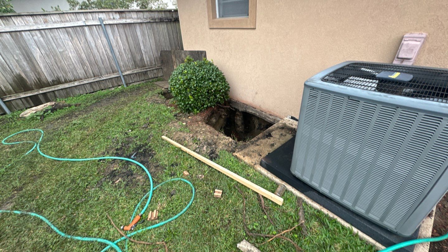 A small yard in Baton Rouge features a hole next to a house, surrounded by a bush, garden hose, bricks, and an air conditioning unit—an essential site for home leveling to ensure the foundation stays secure.