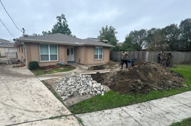 A man stands with a wheelbarrow next to a pile of dirt and rubble in front of a single-story house. The yard is partially dug up, likely addressing common foundation issues often seen in Mississippi homes during construction or landscaping work.