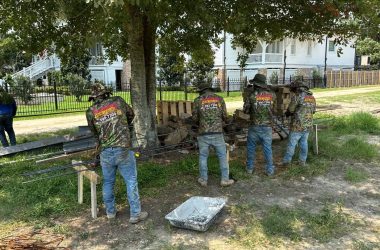 Four workers in camouflage shirts stand under a tree, expertly bending metal bars near a stack of bricks in a grassy area. A person in a blue shirt observes from a distance, ensuring the resources are optimal for the house leveling services being conducted in Metairie.