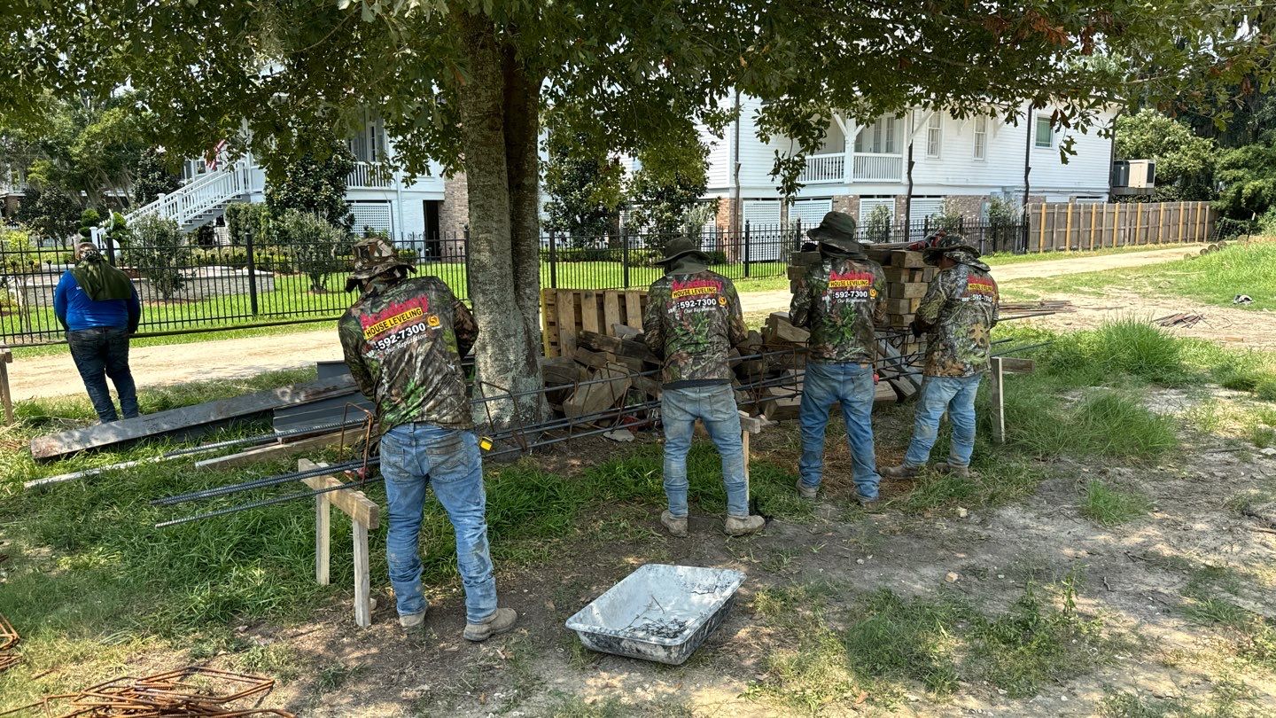 Four workers in camouflage shirts stand under a tree, expertly bending metal bars near a stack of bricks in a grassy area. A person in a blue shirt observes from a distance, ensuring the resources are optimal for the house leveling services being conducted in Metairie.