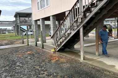A man stands under a raised wooden house by a staircase, reminiscent of the homes in New Orleans. Nearby, there's a muddy area and another elevated house with a walkway. The cloudy sky looms ahead, illustrating the challenges of leveling a pier and beam foundation in this climate.