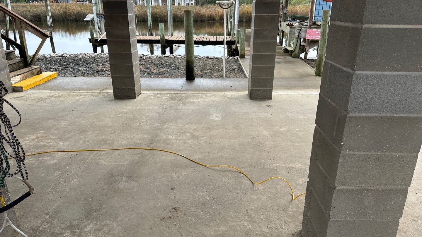 A concrete floor supports a raised structure with visible columns near a waterfront, reminiscent of the classic Pier and Beam Foundation style found in New Orleans. A yellow extension cord snakes across the ground, while wooden docks stand gracefully in the background.