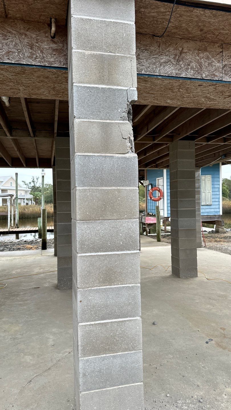 A cracked cinder block pillar supports a raised structure on a concrete slab, reminiscent of the pier and beam foundations often found in New Orleans. Other similar pillars and a lifebuoy can be seen in the background.