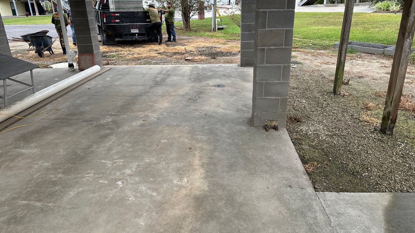 The concrete patio with a partial cinder block structure on the right provides a glimpse into New Orleans' unique architecture. In the background, people and a truck are visible amidst the grass and dirt that frame this ultimate guide to local living spaces.