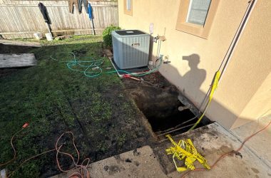 A hole is dug next to a house's exterior wall near an air conditioning unit in New Orleans. A shovel and hose are nearby, hinting at repairs for a sinking foundation, while the shadow of a hard-working figure casts on the wall.