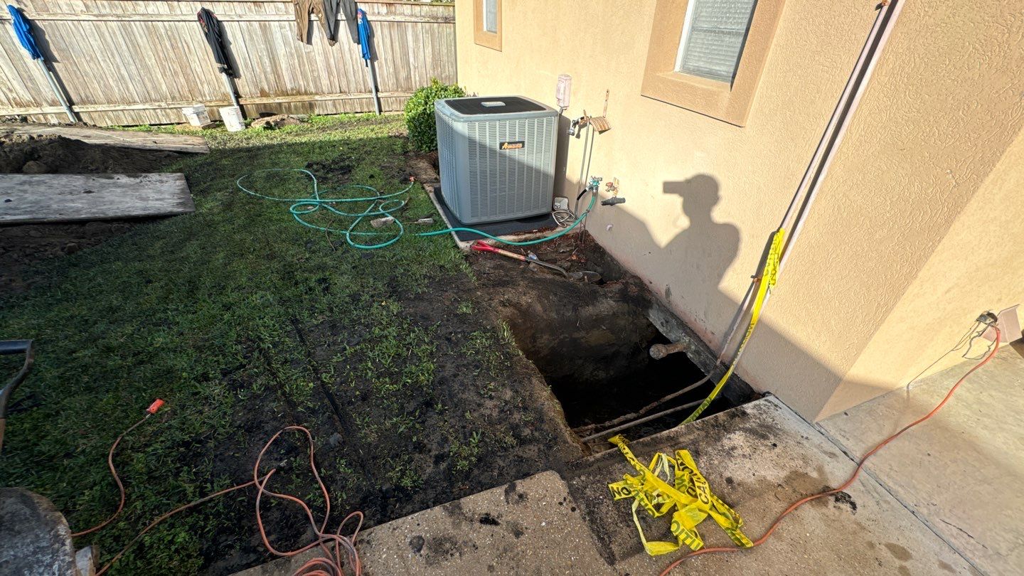 A hole is dug next to a house's exterior wall near an air conditioning unit in New Orleans. A shovel and hose are nearby, hinting at repairs for a sinking foundation, while the shadow of a hard-working figure casts on the wall.