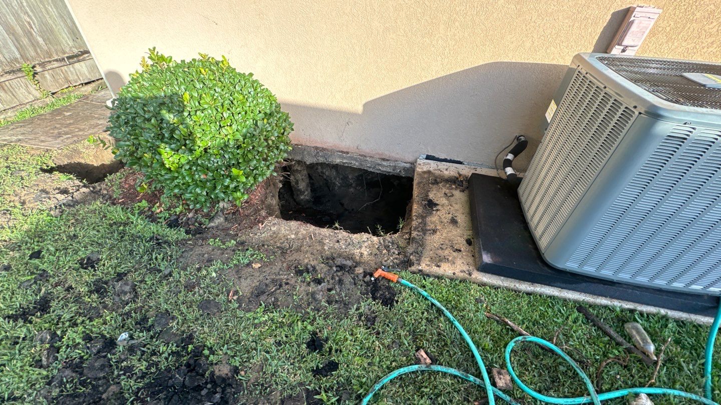A hole next to a building foundation near an air conditioning unit, with a bush and a garden hose on the ground, suggests potential concerns of a sinking foundation, particularly common in areas like New Orleans.